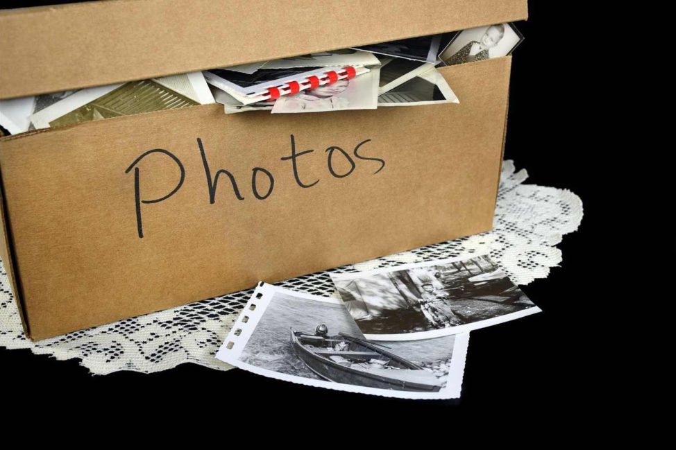 A brown box labeled photos with old photos spilling out of it sits in front of a black background. The box is on top of a lace doily.